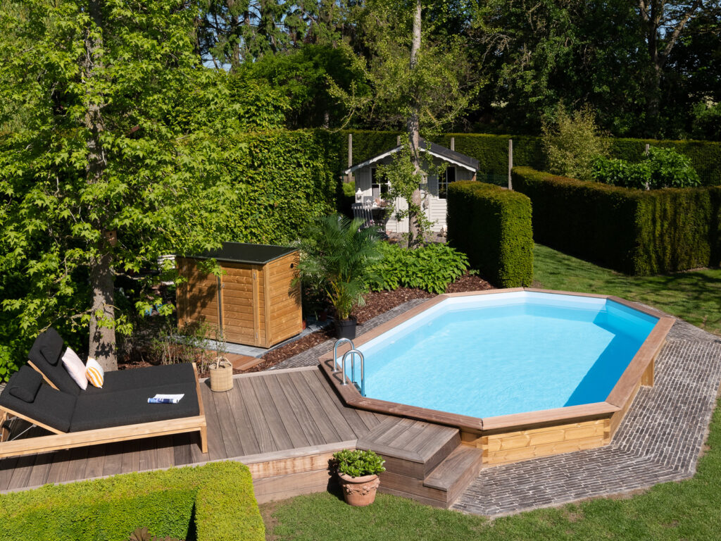 Houten zwembad met terras en ligbed in een groene tuin.