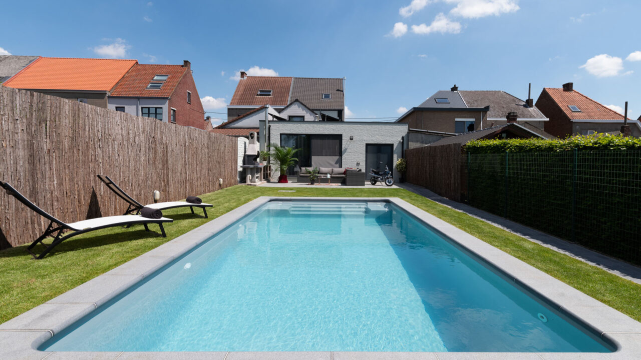 Ingegraven zwembad met grijze liner en natuurstenen terras in een stadstuin, omgeven door gras en ligstoelen.