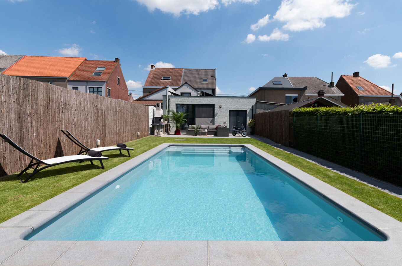 Ingegraven zwembad met grijze liner en natuurstenen terras in een stadstuin, omgeven door gras en ligstoelen.