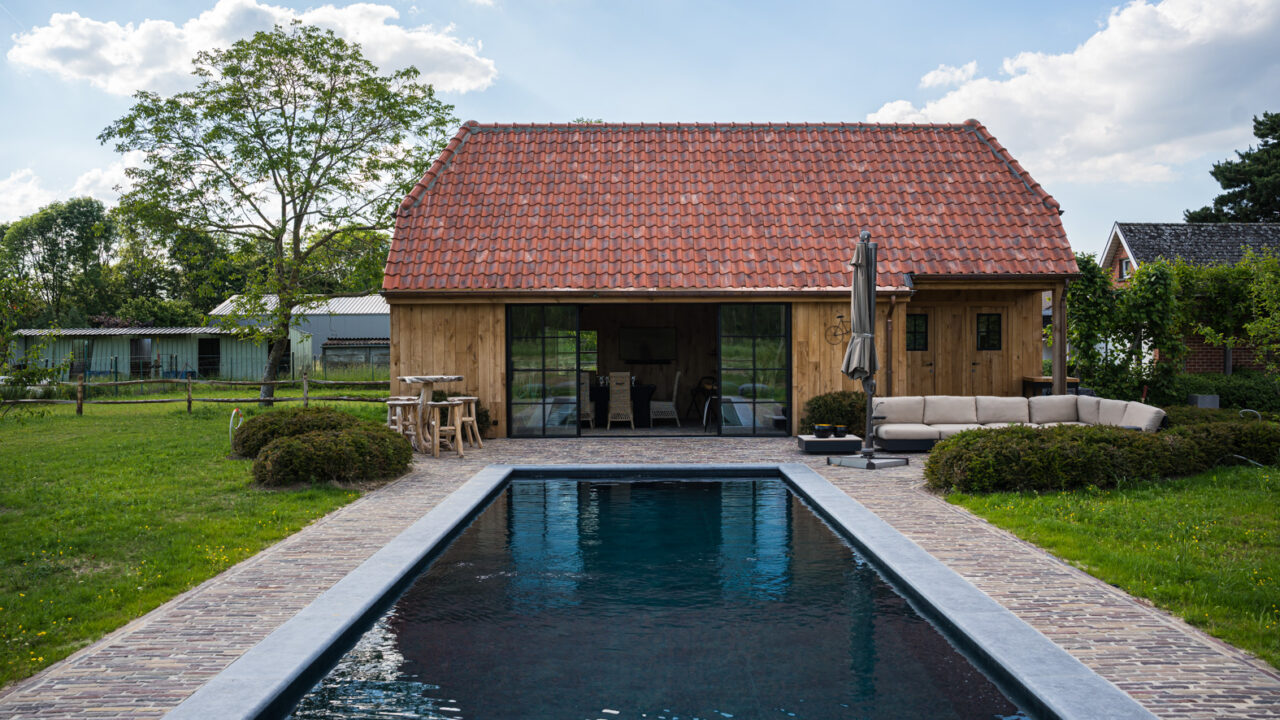 Lang zwembad met donkere waterspiegel voor een poolhouse met rieten dak en houten gevel.